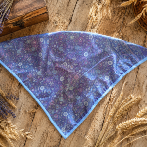 Foulard Manon Lavande déplié sur une table en bois rustique clair. Motif floral dans les tons de violet, bleu et rose, finition biais bleu ciel.